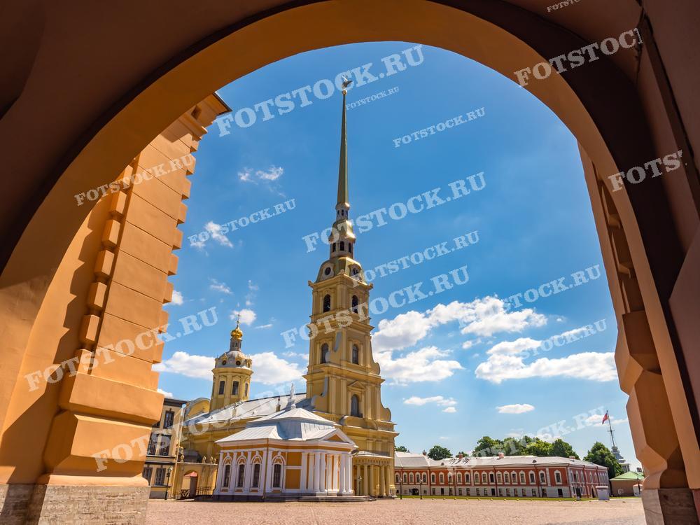 Эта фотография представляет собой вид на Петропавловскую крепость в Санкт-Петербурге, снятый через арку. Яркий летний день подчеркивается глубоким синим небом и белыми облаками, а золотой шпиль крепости выделяется на этом фоне. Изображение подходит для использования в туристических гидах, исторических обзорах и образовательных материалах, а также в качестве постера или иллюстрации в печатных изданиях.